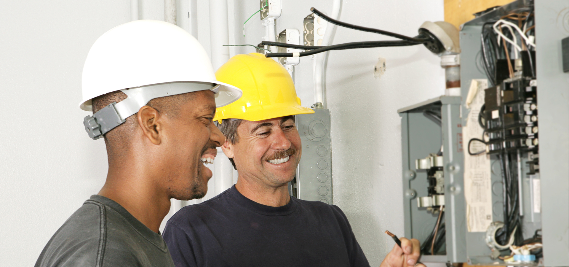 Two men in hard hats inspecting an electrical panel with various switches and meters.
